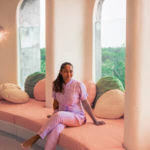 A woman sits relaxed on a long, cushioned bench in front of large arched windows. She is wearing a light purple outfit and is barefoot. The room features soft, pastel colors with pink, green, and white cushions, contributing to a calm and serene atmosphere—an ideal setting for contemplating her Soneva career.