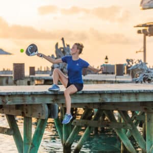 A woman in a blue t-shirt and black shorts is sitting on a wooden dock at Soneva, swinging a paddle racket towards a ball in the air. The dock, part of the Maldives exclusive experience, is situated above water with structures in the background, set against a sunset sky.