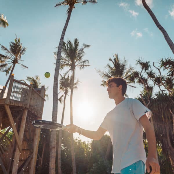 A person in a light t-shirt and blue shorts is playing paddleball outdoors. They are poised to hit the ball with a paddle. Palm trees and a wooden structure are in the background, with the sun shining brightly, creating a vivid, tropical scene reminiscent of Signature Experiences at Soneva Fushi.