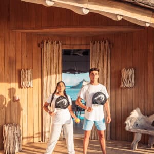 Two people stand confidently on a wooden deck, holding black circular objects against their chests. They wear casual athletic clothing, including white shirts. This Maldives experience on the Soneva wooden structure offers a stunning view of blue skies and water in the background.