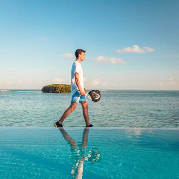 A person dressed in a white shirt and blue shorts walks on the edge of an infinity pool, holding a tennis racket, with calm ocean water and a small green island in the background under a clear blue sky. This scene captures the essence of a Maldives Exclusive Experience at Soneva.