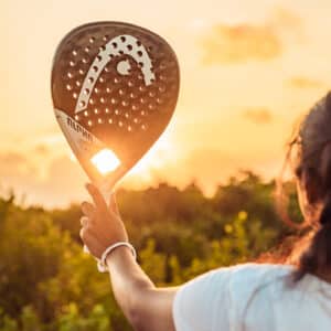 A person holding a paddle racket up to the sky with the sunset in the background, reminiscent of a Maldives Exclusive Experience. The racket casts a shadow, framing the setting sun. The person is wearing a white shirt and bracelet, their hair tied up. Greenery surrounds this picturesque scene at Soneva.