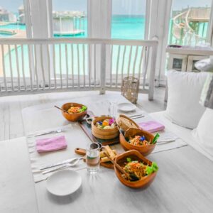 A set table with wooden bowls of colorful salad and a bamboo steamer filled with dumplings. Pink napkins, cutlery, a water glass, and condiments are arranged on a white table, overlooking turquoise water and overwater bungalows. Soneva Exclusive Offers enhance this breathtaking dining experience.