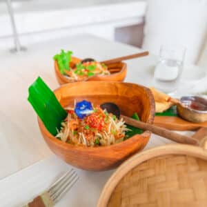 A vibrant meal served in wooden bowls, featuring a fresh salad garnished with a blue edible flower and green leaves, evokes the Soneva Maldives luxury. The table showcases a bamboo basket and dipping sauces, set up for dining with a fork, napkin, and a glass of water in the background.