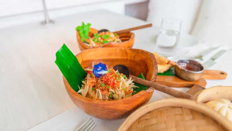 A vibrant meal served in wooden bowls, featuring a fresh salad garnished with a blue edible flower and green leaves, evokes the Soneva Maldives luxury. The table showcases a bamboo basket and dipping sauces, set up for dining with a fork, napkin, and a glass of water in the background.