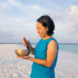 A woman in a blue sleeveless top stands on a sandy beach with clear skies, holding and striking a singing bowl with a mallet. The ocean and a few clouds are visible in the background. She appears to be meditating or enjoying the serene environment, reminiscent of Soneva Exclusive Offers.