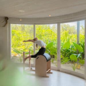 A person is doing a backbend stretch on Pilates equipment next to a large panoramic window in a bright room. Lush greenery outside adds a serene and nature-infused backdrop to the scene, reminiscent of Soneva Exclusive Offers. The room has light wood floors and a high ceiling.