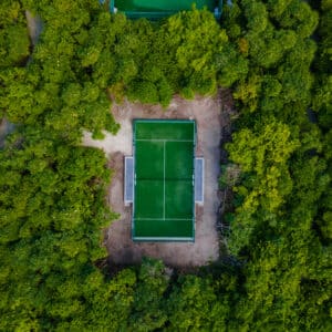 Aerial view of a rectangular green tennis court with white lines, surrounded by dense, lush trees. The vibrant greenery envelops the court, creating a stark contrast between the natural environment and the man-made structure—a true Soneva Exclusive Experience Maldives moment.