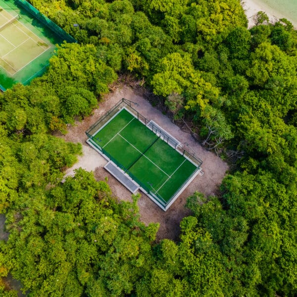 Aerial view of a tennis court nestled in lush green forest, offering an exclusive experience reminiscent of the Maldives. The court, surrounded by dense trees, appears isolated and serene. Another tennis court is partially visible at the top left of the image. A dirt path leads to the main court, evoking Soneva luxury.