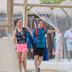 A woman and a man, both dressed in sportswear and carrying bags, walk through a netted area in an outdoor setting with greenery and wooden structures at Soneva, offering the perfect backdrop for a Maldives exclusive experience. The woman wears a black tank top and pink shorts, while the man sports a blue shirt and black shorts.