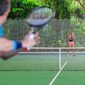 Two people, one in the foreground and one in the background, are playing tennis on an outdoor court surrounded by greenery. The person in the foreground, seen from behind, is about to hit the ball, while the person in the background prepares to return the shot—an exclusive experience reminiscent of Soneva's serene Maldives setting.