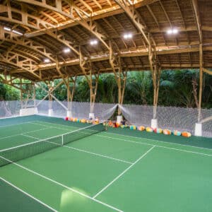 An indoor tennis court with a wooden arched roof set against a backdrop of lush greenery exudes the Soneva Exclusive Experience Maldives. Two players are engaged in a game; one is serving while the other is ready to receive. Brightly colored balls and equipment are lined up along the side.