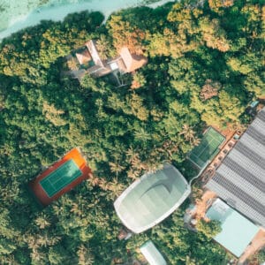 Aerial view of a lush, tropical area featuring various structures including a tennis court, a large dome-shaped building, and additional buildings nestled among dense greenery, with clear turquoise waters visible at the top of the image—a true Soneva Exclusive Experience Maldives.