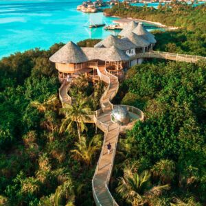 Aerial view of a tropical resort with thatched-roof huts connected by winding wooden walkways surrounded by lush greenery. Clear turquoise waters border the resort, with a line of similar huts stretching into the distance under a bright blue sky, highlighting Soneva Exclusive Offers.