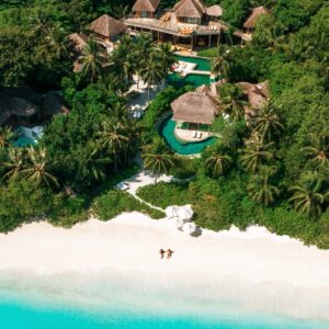 Aerial view of a tropical beach with turquoise water and white sand. Two people lie on the sand near the shoreline. Behind the beach, lush green vegetation surrounds luxury villas with thatched roofs and swimming pools. Palm trees dot the landscape, offering Soneva Exclusive Offers to visitors.