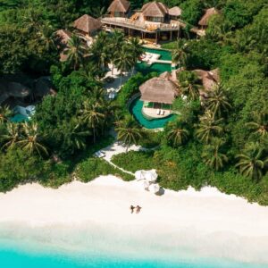 Aerial view of a tropical beach resort surrounded by lush greenery. The resort features multiple thatched-roof buildings, winding pools, and palm trees. Two people relax on the pristine, white sand beach near the turquoise ocean under a white umbrella, enjoying Soneva Exclusive Offers.