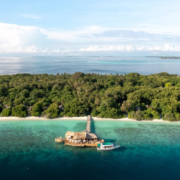 Aerial view of a tropical island with dense forest and a sandy beach. A large thatched-roof building on a pier extends into the clear blue ocean, with a boat docked alongside it. The horizon shows a calm sea meeting a cloud-dotted sky, offering an idyllic backdrop for Soneva Exclusive Offers.