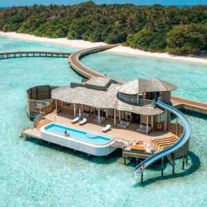 A luxurious overwater villa with a private pool, slide, and sunbathing deck sits on crystal-clear turquoise water. A winding wooden walkway connects the villa to a lush, green island. The scene conveys a tropical paradise with clear blue skies and is part of the Soneva Exclusive Offers.