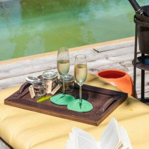 A poolside setting with a tray holding two champagne flutes, corks, and small jars on a yellow sunbed. Nearby is an open book and an ice bucket with a bottle of champagne, evoking the allure of Soneva Exclusive Offers. The pool and lush greenery are visible in the background.