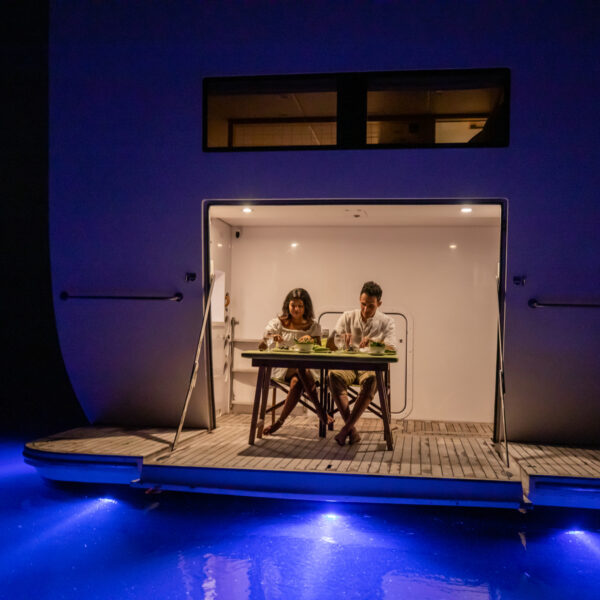 A couple enjoys dinner on the deck of a luxury yacht illuminated by blue underwater lights at night. They are sitting in a cozy, open area with a modern, minimalist design, near the calm waters of the Maldives, giving a serene and romantic atmosphere perfect for a holiday escape.