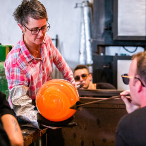 A person with short hair and glasses, wearing a plaid shirt and protective sleeve, holds a large, glowing orange glass piece at the end of a metal rod. Another person in glasses uses a tool to shape it while a third observer watches, reminiscent of the craftsmanship seen in Maldives Exclusive Experience by Soneva.