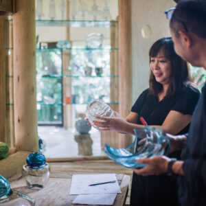 A woman and a man are examining glass art pieces in a well-lit room. The woman is holding a clear glass bowl, while the man is holding a blue curved glass piece. Various glass artworks are displayed on a table and shelves in the background, evoking the exclusivity of a Maldives Soneva experience as they discuss the items.