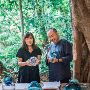 Two people stand in a lush, outdoor setting surrounded by large trees. They are both smiling and holding colorful glass sculptures. The table in front of them displays more glass art pieces and some papers, evoking the creative vibe akin to a Maldives Exclusive Experience at Soneva.