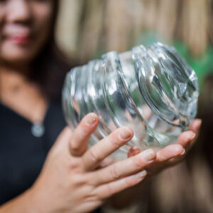 A person holding a broken glass jar with both hands, sharp, jagged edges where it has shattered. The background is blurred, drawing attention to the jar and the person&#039;s hands with light-colored nail polish—a stark contrast to the serene beauty of a Maldives exclusive experience at Soneva.