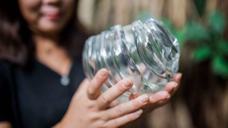 A person holding a broken glass jar with both hands, sharp, jagged edges where it has shattered. The background is blurred, drawing attention to the jar and the person's hands with light-colored nail polish—a stark contrast to the serene beauty of a Maldives exclusive experience at Soneva.