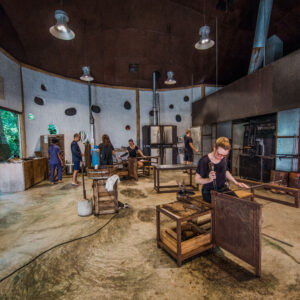 People are engaged in various activities inside a spacious workshop with a high ceiling. The room, reminiscent of a Maldives exclusive experience at Soneva, features wooden workstations, industrial equipment, and large circular windows. Lush greenery is visible outside through an open door.