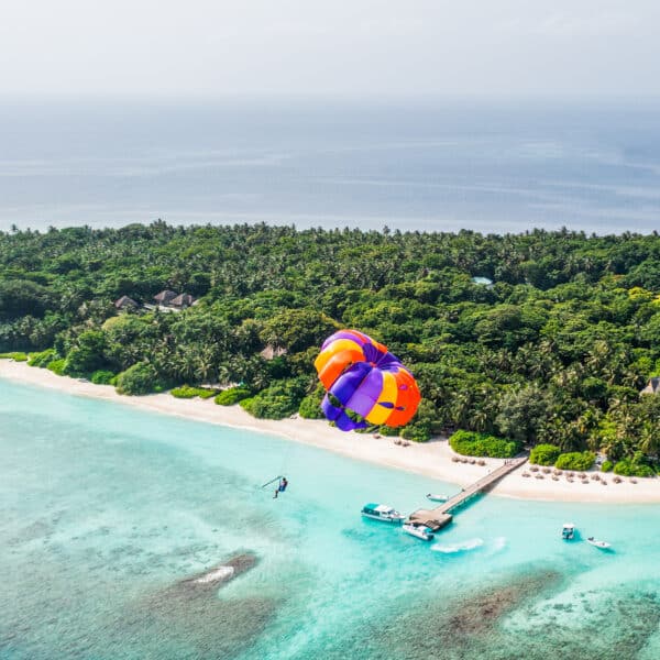 A vibrant orange and purple parasail soars over turquoise waters and a lush green island with a white sandy beach, part of the Soneva Exclusive Experience Maldives. Boats dock at a pier, while beach chairs line up near the shoreline, all beneath clear skies that enhance this scenic tropical destination.