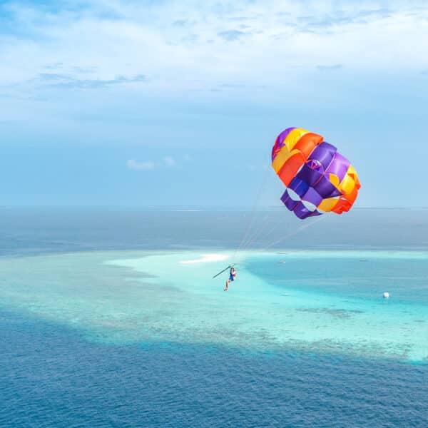 A person parasails over a turquoise ocean near a small, sandy island, capturing the essence of a Soneva Exclusive Experience Maldives. The vibrant parasail features red, orange, and purple colors. The clear water and distant horizon create a tranquil backdrop under a partly cloudy sky.