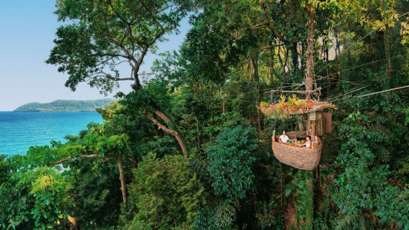 A group of people dine in a pod-shaped wicker structure suspended among trees in a lush forest, with a view of the ocean in the background. This exclusive experience at Soneva in Thailand offers serene surroundings, dense greenery, and clear blue waters in the distance.