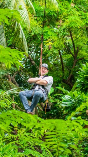 A person wearing a helmet and harness glides through a lush, green forest canopy on a zipline, smiling. The background is filled with vibrant leaves and dense foliage, offering an adventure reminiscent of a Soneva Exclusive Experience in the Maldives.