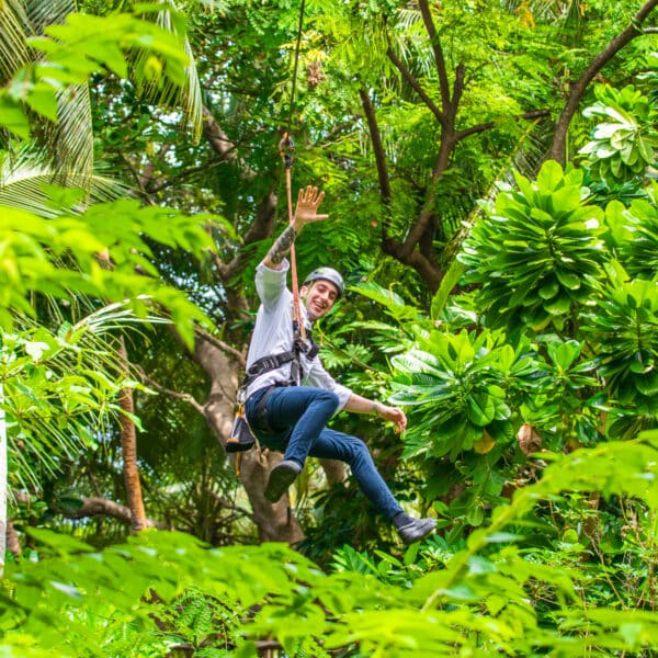 A person wearing a helmet and harness is zip-lining through a lush green jungle, enjoying what feels like a Soneva Exclusive Experience Maldives adventure. Their left hand grips the pulley while their right hand waves joyfully. Bright green leaves surround them, enhancing the vibrant and adventurous atmosphere.