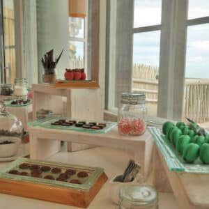 A dessert table set by a window displays a variety of treats. There are glass jars and plates filled with assorted chocolates, macarons, and candies. Wooden stands hold the desserts at different levels, creating an inviting and elegant presentation reminiscent of the indulgent spreads at a Maldives luxury resort like Soneva.