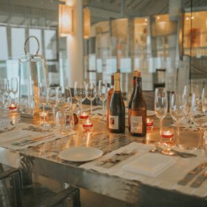 A dining table at the Maldives Luxury Resort Soneva is elegantly set with wine glasses, candles, and several bottles of fine wine. The tableware is neatly arranged with plates, forks, knives, and napkins. The background reveals a modern, well-lit interior with large windows showcasing the resort’s stunning views.