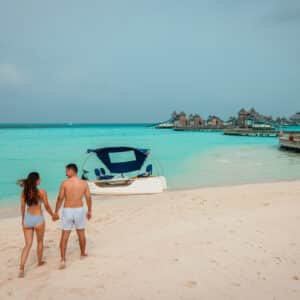 A couple walks hand in hand on a pristine sandy beach towards a small boat with a blue canopy. The turquoise ocean stretches out, meeting the horizon. Overwater bungalows are visible in the distance under a clear sky, as they revel in their Soneva Exclusive Offers getaway.