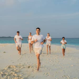 A group of five people jogging on a sandy beach near turquoise ocean waters. They are all dressed in white and light-colored athletic wear, running barefoot on the shore. The sky is mostly clear with a hint of clouds, embodying the tranquility captured in Soneva Exclusive Offers.