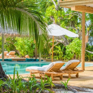 A serene tropical poolside scene with two wooden lounge chairs and white cushions on a wooden deck. A large white umbrella provides shade, surrounded by lush green plants and trees. A hammock hangs in the background, and there is a calm pool reflecting the greenery—truly a Soneva Exclusive Offers experience.