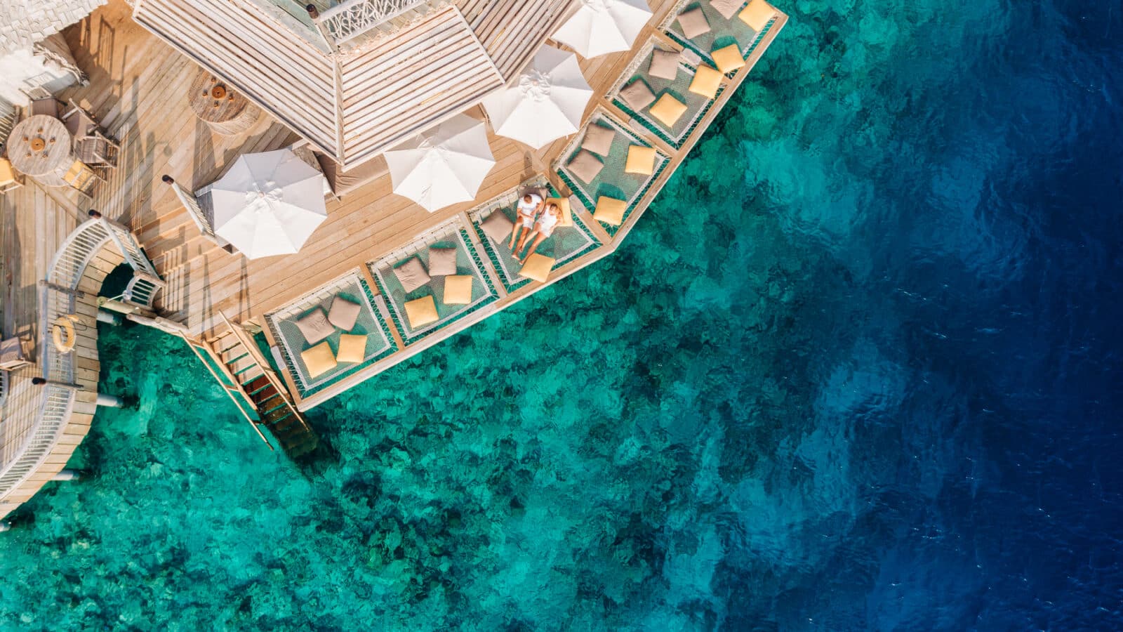 Aerial view of a luxurious seaside resort featuring several sun loungers and umbrellas on wooden decks that extend over an expanse of clear turquoise and deep blue water. A couple relaxes on one of the loungers, enjoying the serene and scenic coastal setting with exclusive Soneva offers.