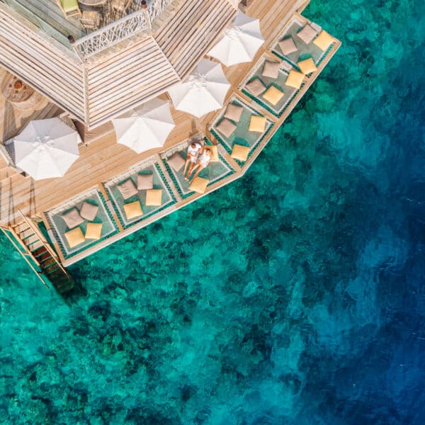 Aerial view of a luxurious seaside resort featuring several sun loungers and umbrellas on wooden decks that extend over an expanse of clear turquoise and deep blue water. A couple relaxes on one of the loungers, enjoying the serene and scenic coastal setting with exclusive Soneva offers.