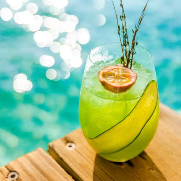 A refreshing green cocktail sits on a wooden deck by the water at Soneva Maldives. The drink is garnished with a cucumber slice, half a passion fruit, and a small sprig of herbs. The sparkling turquoise water in the background glistens in the sunlight, enhancing the luxury experience.