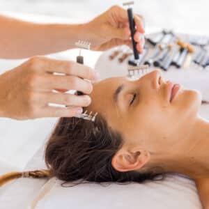 A person with closed eyes is lying on a white towel, receiving a luxurious facial treatment. Two hands are seen holding small, comb-like tools above the person's forehead and temples. The background is blurred, focusing attention on the Soneva Exclusive Offers experience.