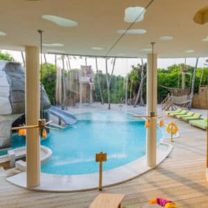 A luxurious indoor-outdoor pool area perfect for a family holiday, featuring a rock-shaped slide, multiple lounging areas with green cushions, and large wooden elements. Sunlight streams through ceiling cutouts, and the surroundings are lush with greenery and tall palm trees—reminiscent of Soneva in the Maldives.