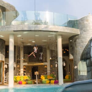 A person in motion is zip-lining into the pool area of a luxurious resort, perfect for a family holiday at Soneva in the Maldives, featuring a stone building with multiple levels, large glass windows, and colorful seating arrangements. The setting is modern and playful, with an elephant-themed structure to the right.