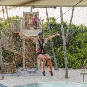 A person wearing a helmet and safety harness is zip-lining over a pool, with arms outstretched. Water droplets splash everywhere as they descend. In the background, a spiral staircase leads up to the zip-lining platform surrounded by lush greenery at Soneva in the Maldives, perfect for a family holiday.