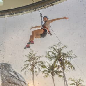 A person wearing a helmet and safety harness is airborne, swinging through a ring of water against a backdrop of palm trees and a cloudy sky during their family holiday at Soneva in the Maldives. They are holding onto a rope with one hand, legs spread outward and free arm extended, appearing mid-flight.