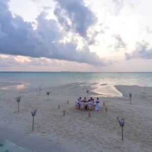 A group of people dining at a single table set up on a sandy beach at a Maldives luxury resort. The table is surrounded by tiki torches, and the ocean with calm waves is visible in the background under a cloudy sky, creating an unforgettable Soneva food experience.