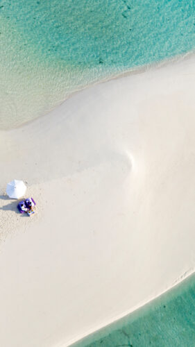 Aerial view of a sandbar surrounded by clear turquoise water at a Maldives luxury resort. Two people sit under a white umbrella on the sandbar's center, casting a shadow. The smooth white sand tapers off into the shallow waters around it, offering an idyllic setting reminiscent of Soneva's allure.